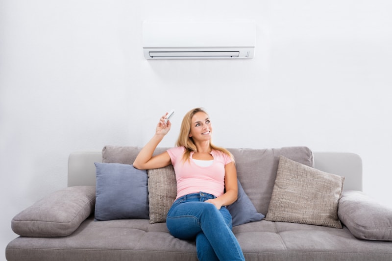 Ductless ACs Improve Indoor Air Quality and Control Humidity. Young Happy Woman Sitting On Couch Operating Air Conditioner With Remote Control At Home.