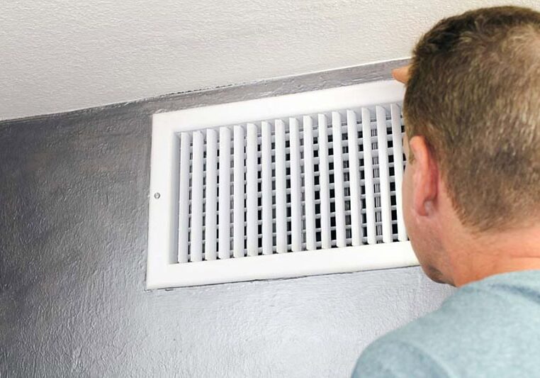 Mature male peering inside an upper wall white grid air duct on a silver wall near a white ceiling. A guy inspecting a heating and cooling air register duct for maintenance.