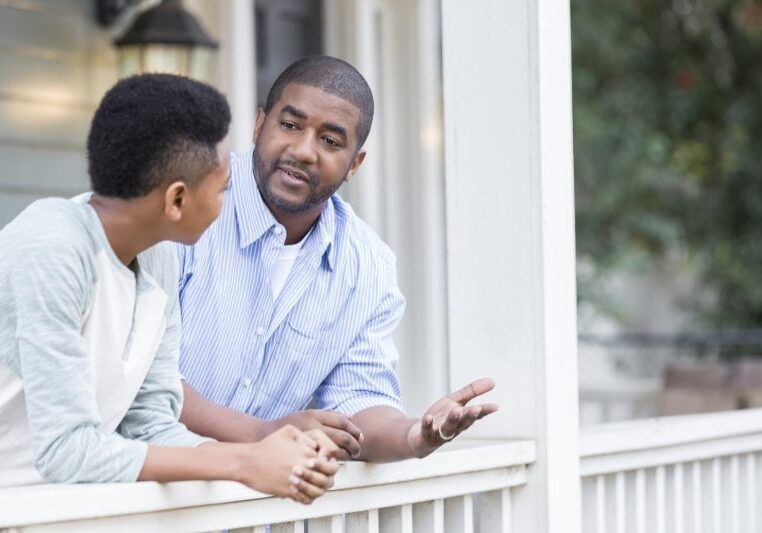 A father talks with his hands as he leans against the railing of his front porch with his preteen son and has a serious discussion.