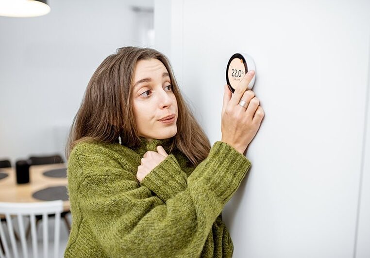 woman shivering in sweater as she adjusts her thermostat at home.